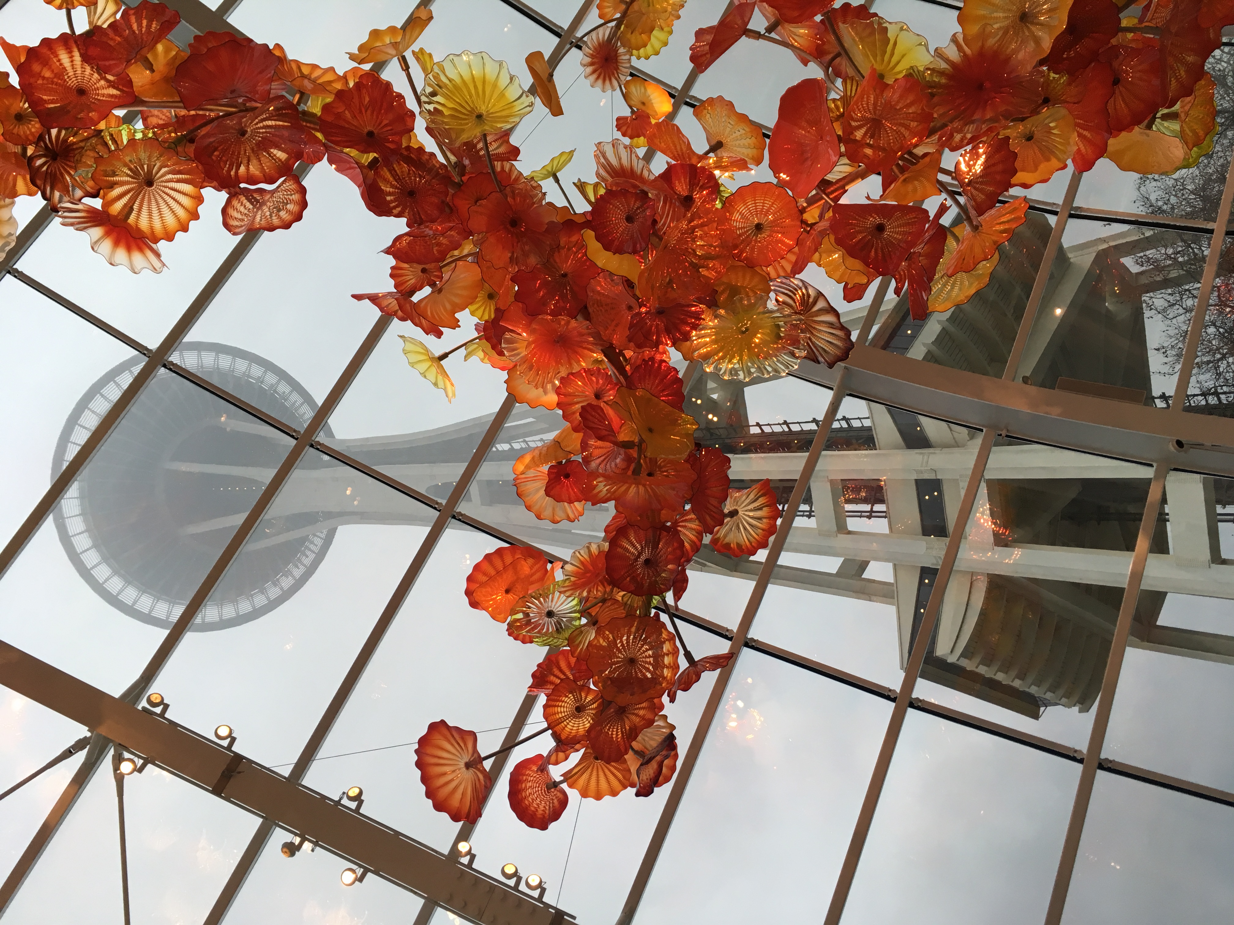 Looking up from inside a building with a glass roof, there are orange blown glass flowers in the foreground with the Seattle Space Needle in the the foggy background.