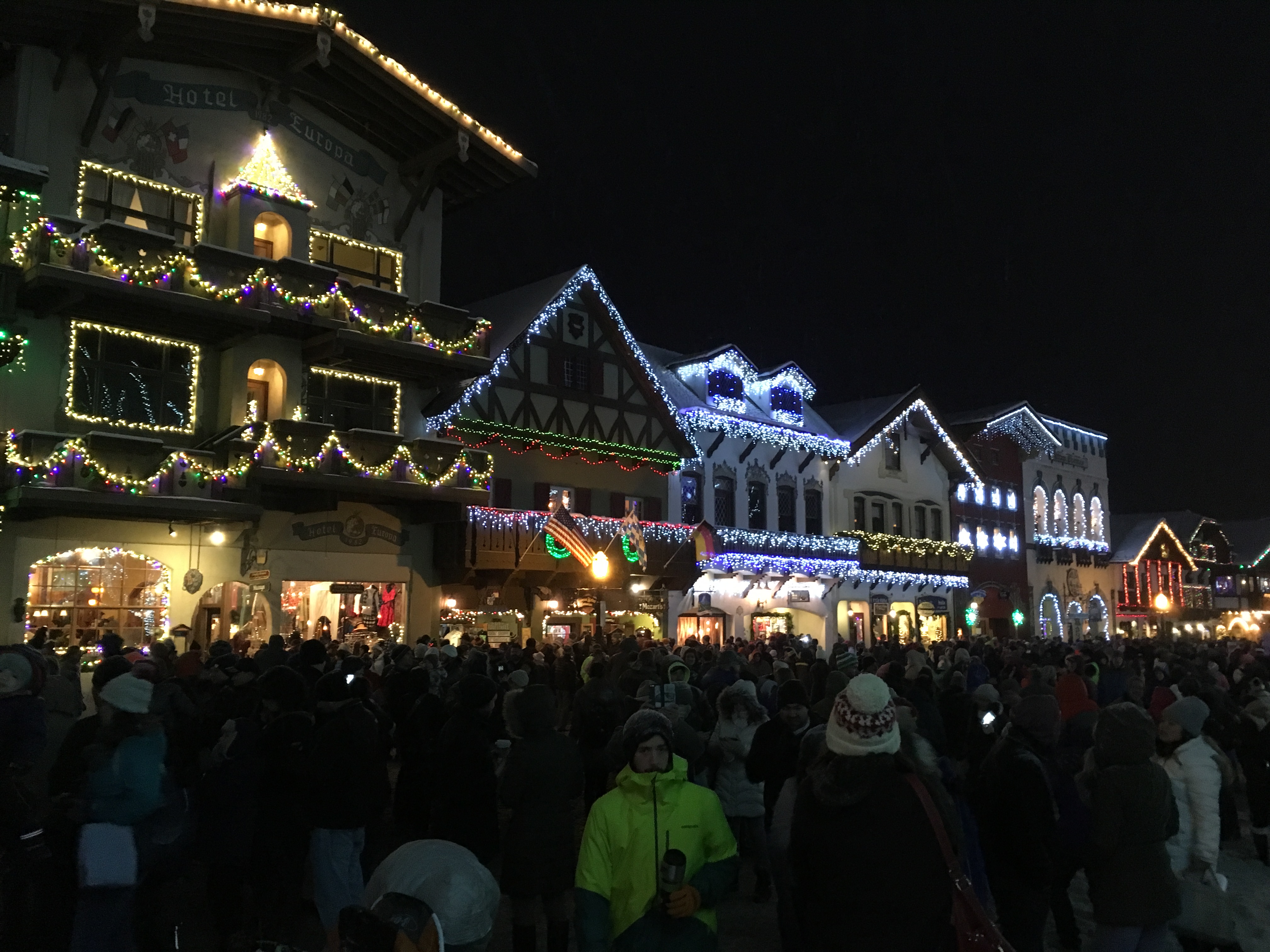 A bavarian village at night with rooftops lined with Christmas lights. The street in the foreground is completely full of people.