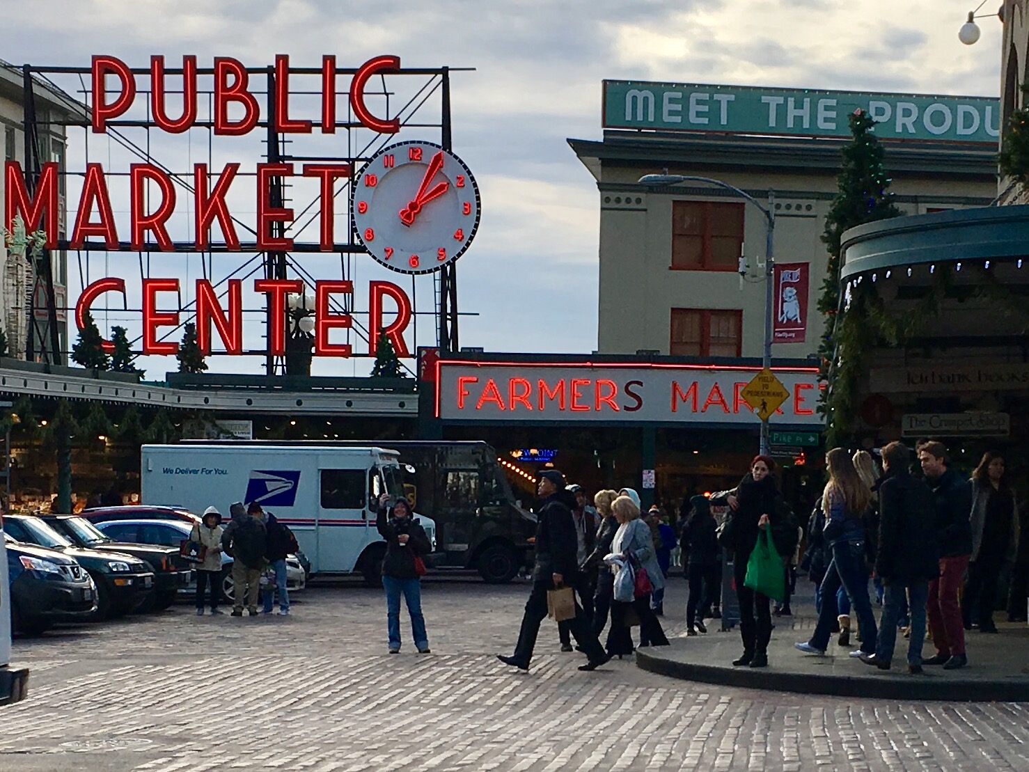 The neon signs reading 'Public Market Center' and 'Farmers Market' in front of Pike Place Market, with people in the foreground waiting to cross the street.